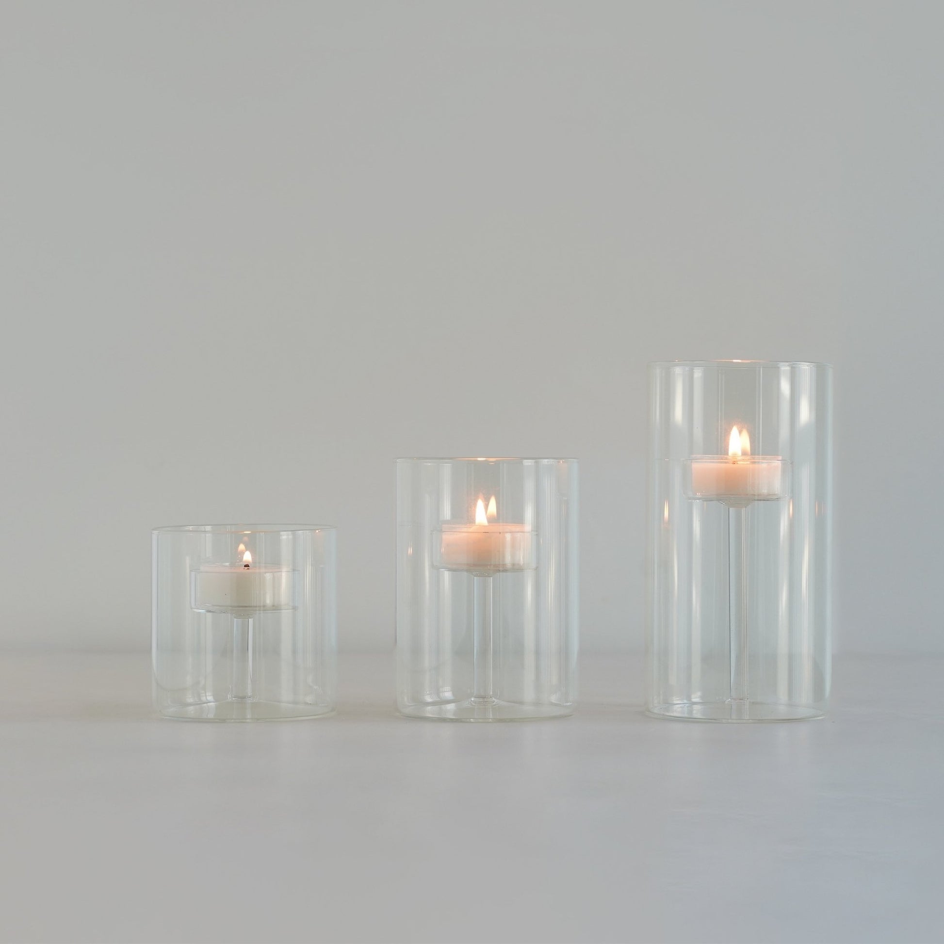 연한 배경 위에 줄지어 놓인 투명 유리 실린더 캔들 홀더와 안에서 따뜻하게 켜진 티라이트 캔들이 보이는 미니멀 제품 연출 사진