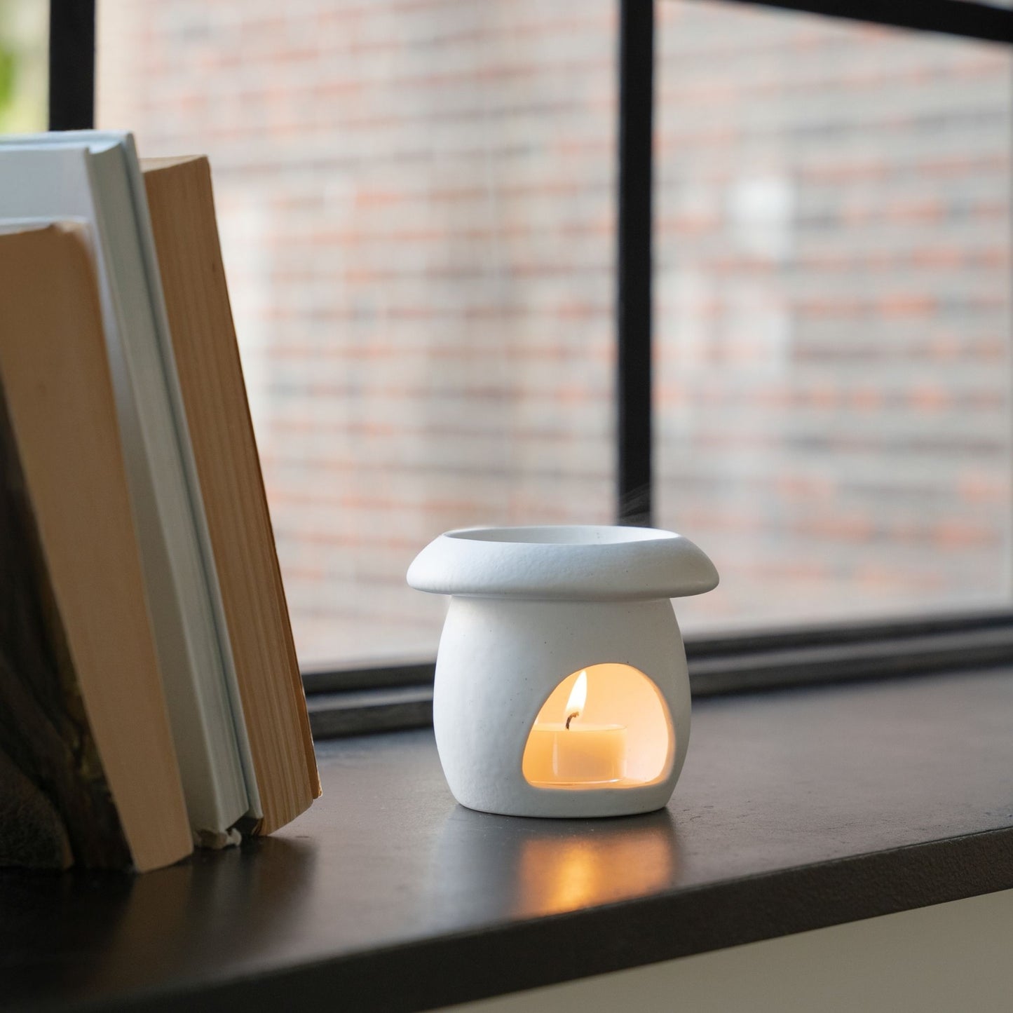 창가 검은 창틀 위 책들 옆에 놓인 화이트 머쉬룸 오일버너와 안에서 타고 있는 티라이트 캔들, 창밖 벽돌 건물이 배경인 사진
