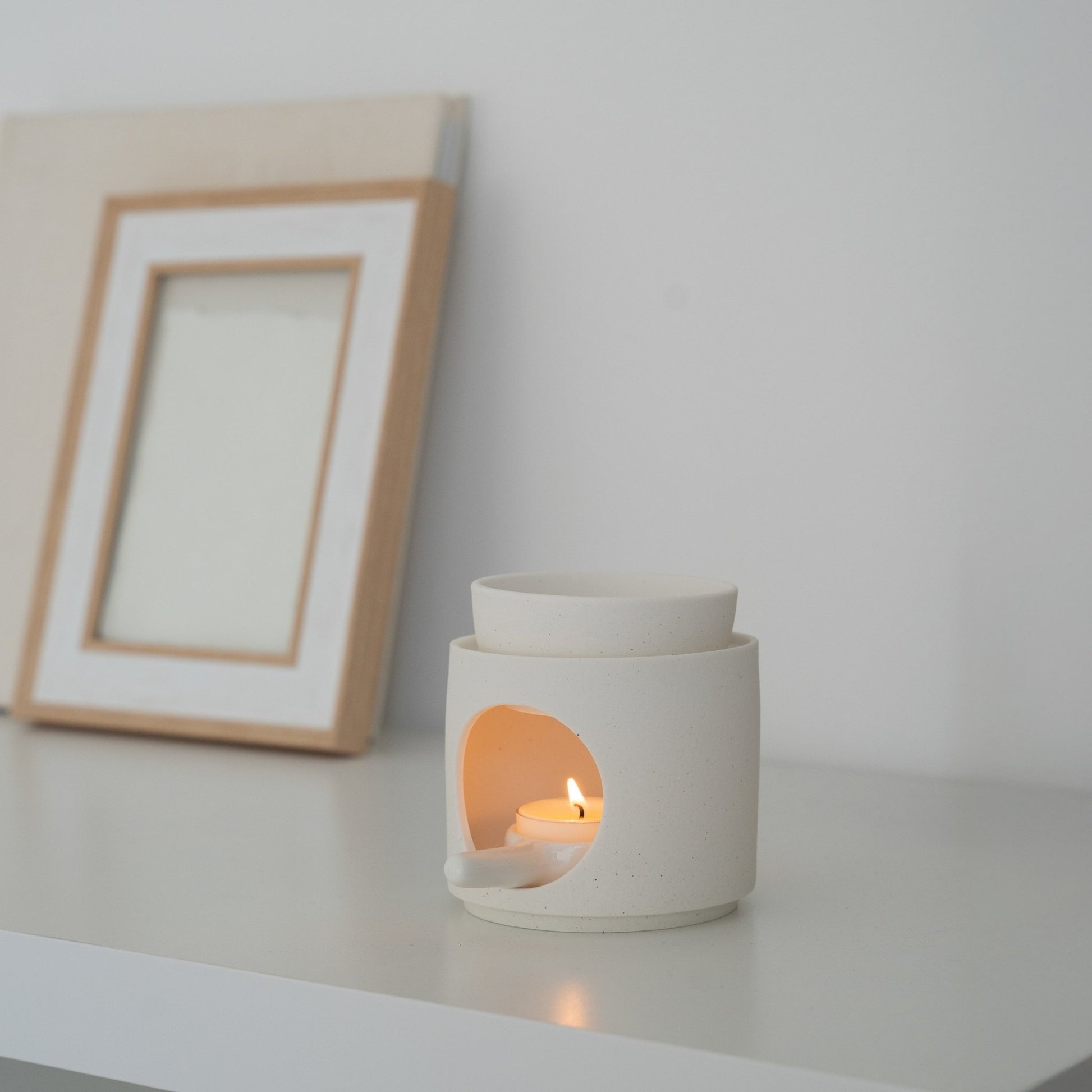 화이트 선반 위 빈 액자 옆에 놓인 화이트 세라믹 오일버너와 은은한 불빛의 티라이트 캔들이 보이는 연출 사진