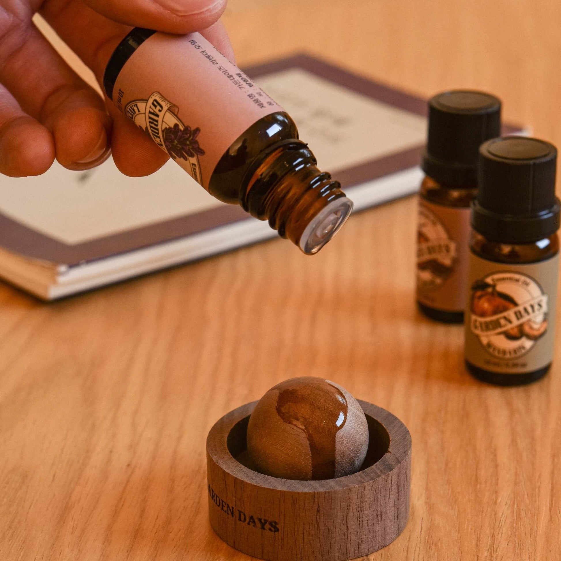 노트와 에센셜오일 병 옆 책상 위에서 호두 브리즈볼 우드 발향 제품 위로 가든데이즈 천연 100% 에센셜오일을 떨어뜨리는 클로즈업 사진