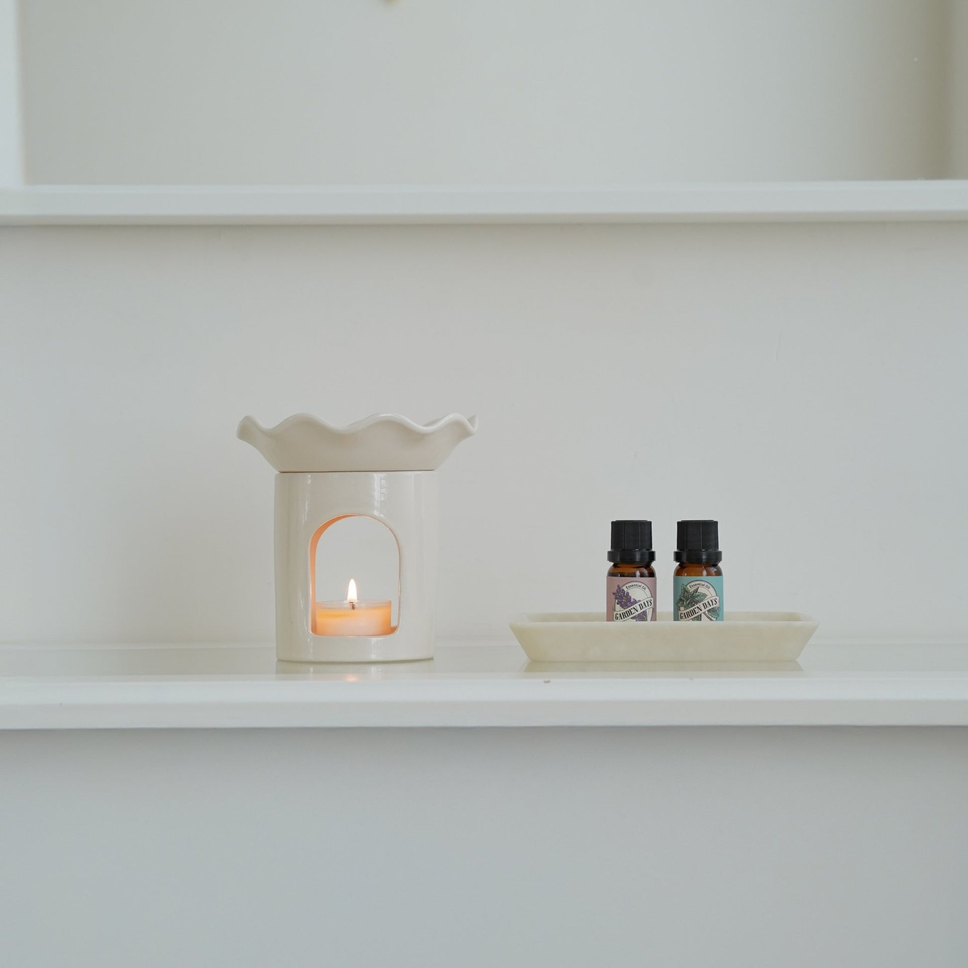 화이트 벽 선반 위에 놓인 웨이브 모양 화이트 오일버너와 티라이트 캔들, 가든데이즈 에센셜오일 병 두 개가 함께 보이는 미니멀 인테리어 사진
