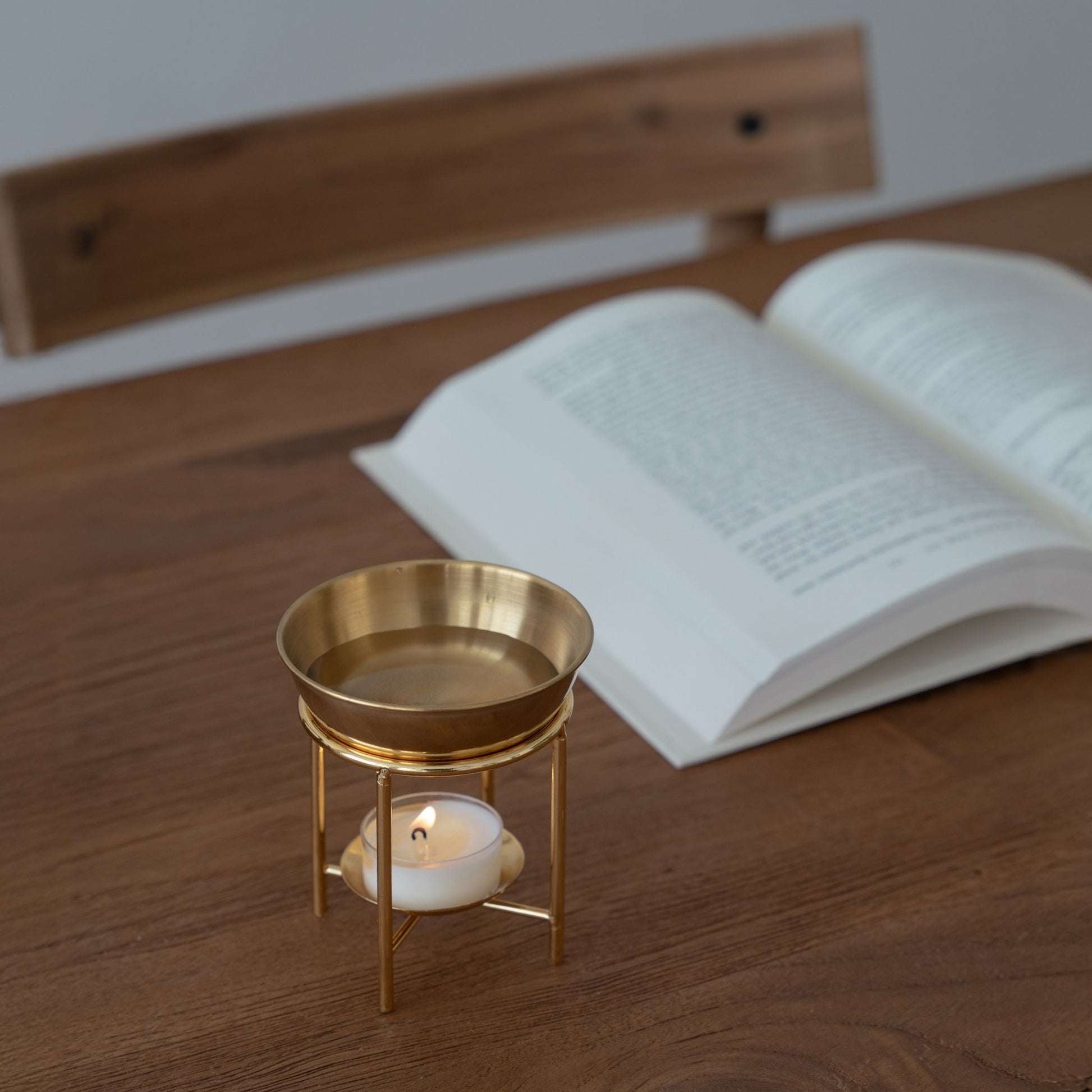 나무 테이블 위 펼쳐진 책 옆에 놓인 골드 스탠드 오일버너와 안에서 켜진 티라이트 캔들이 보이는 독서 연출 사진
