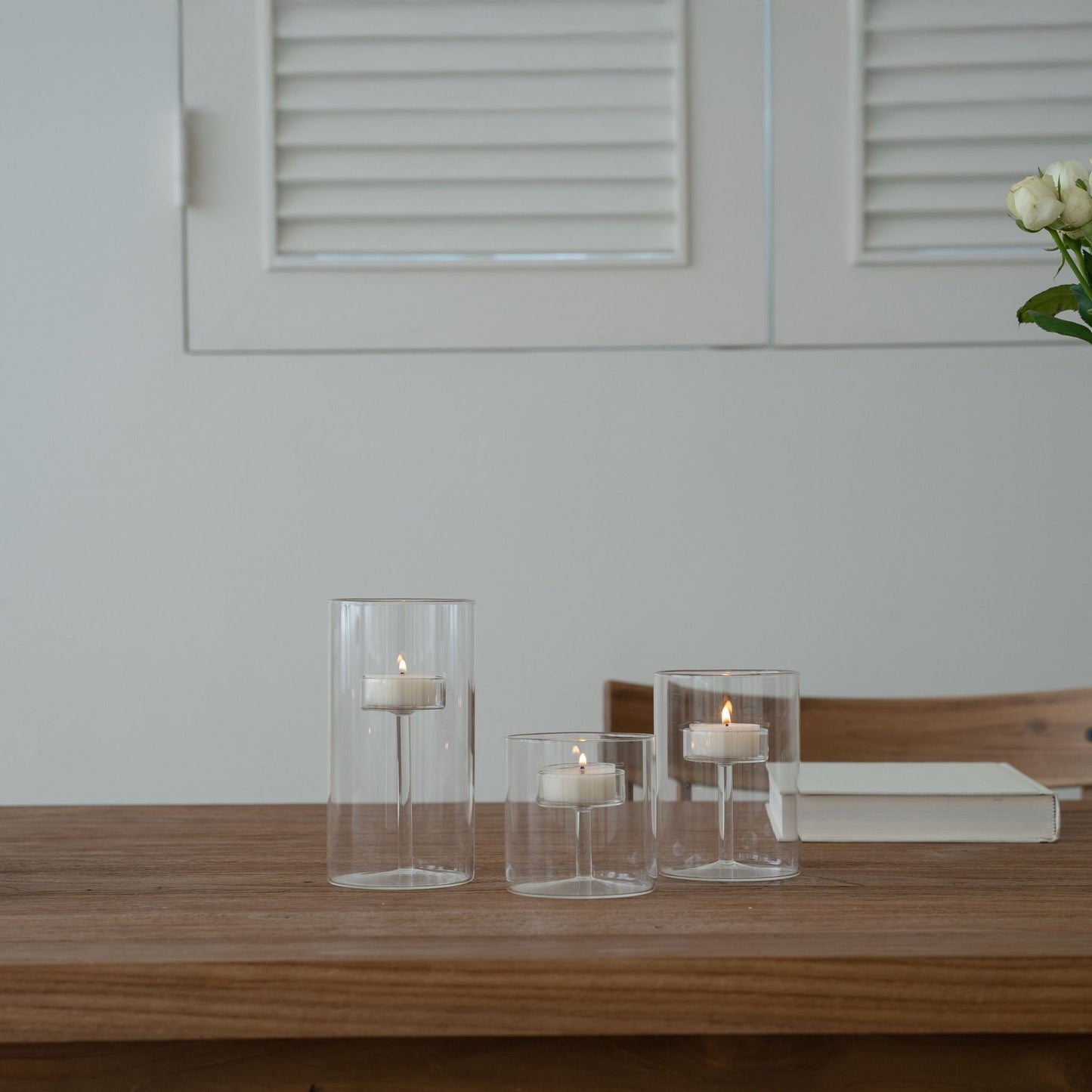 나무 테이블 위 책과 함께 놓인 투명 유리 실린더 캔들 홀더 세트와 불이 켜진 티라이트 캔들이 보이는 거실 연출 사진