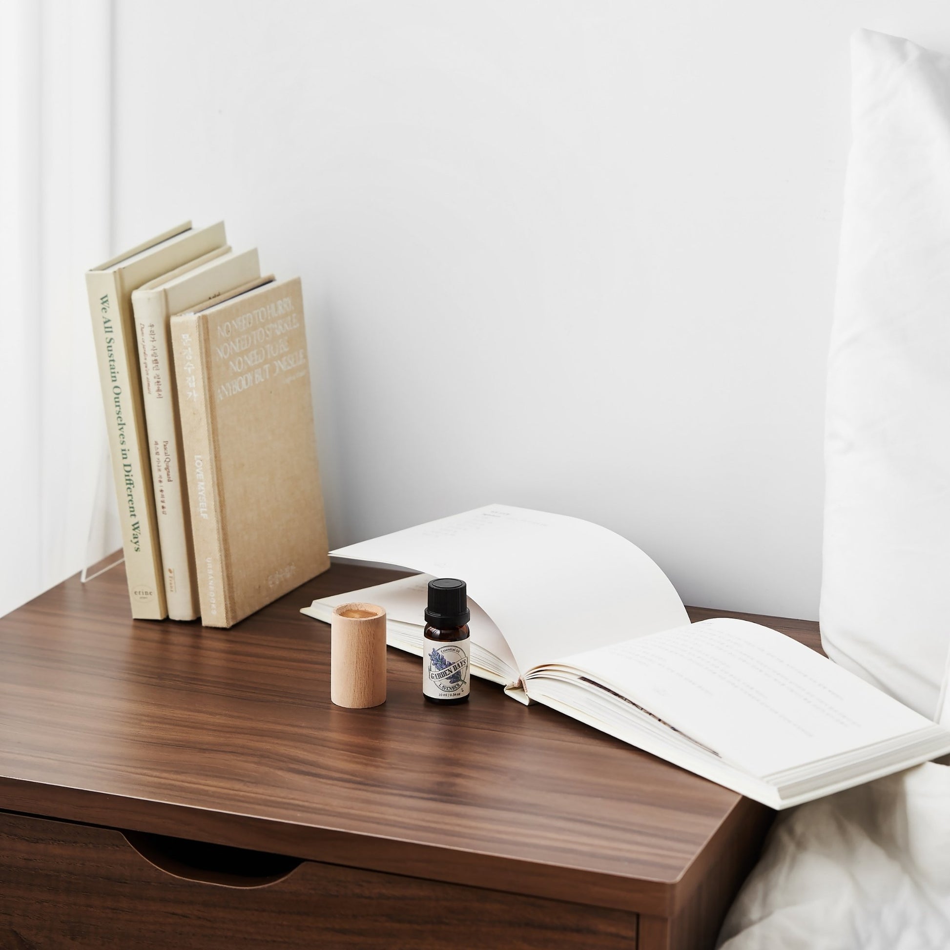 침대 옆 나무 협탁 위 책 더미와 펼쳐진 책 옆에 놓인 너도밤나무 원목 실린더 발향 우드가 보이는 아늑한 침실 연출 사진