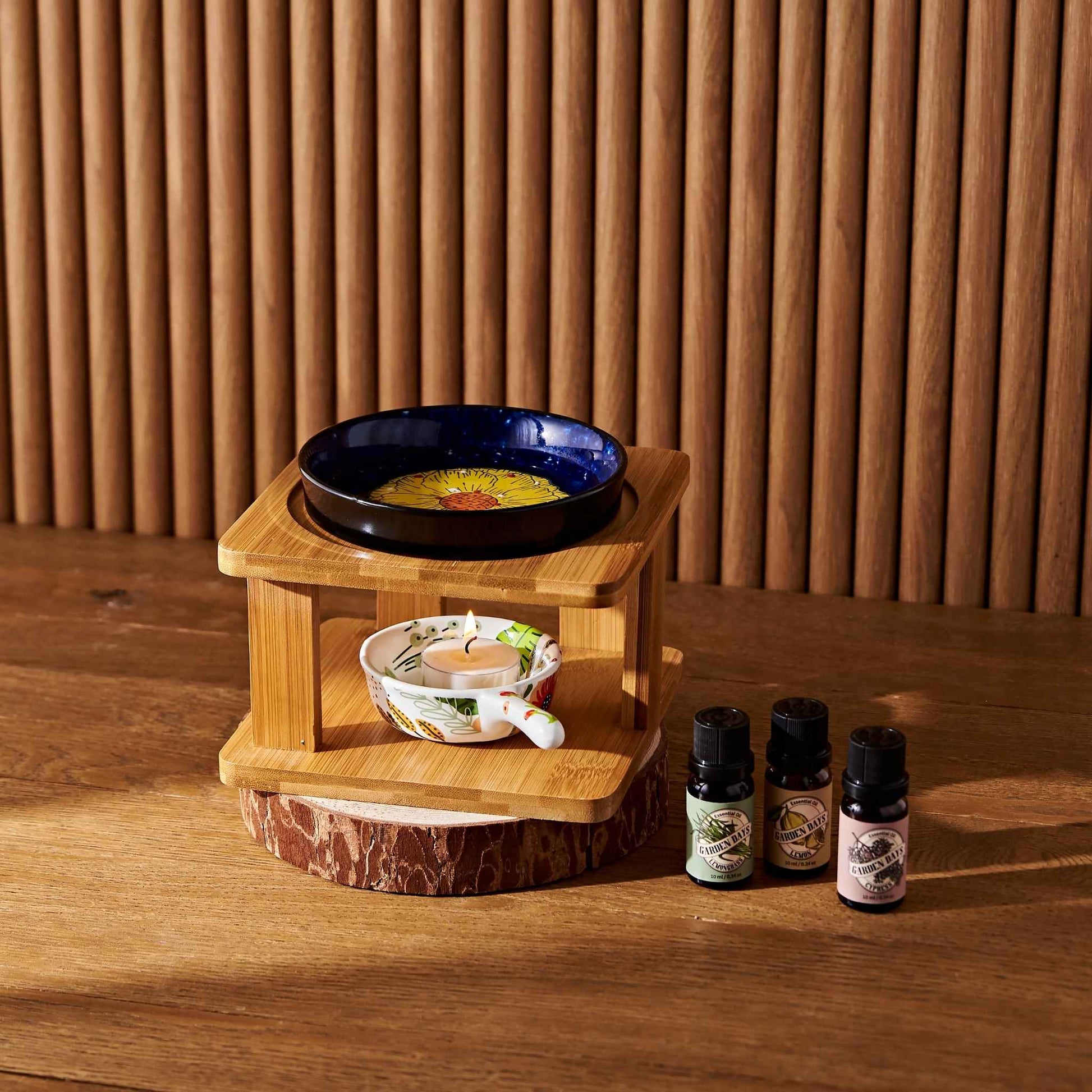 나무 테이블 위 우드 스탠드 오일버너와 네이비세라믹 볼, 아래 티라이트 캔들, 앞에 놓인 가든데이즈 에센셜오일 병이 보이는 따뜻한 분위기의 연출 사진