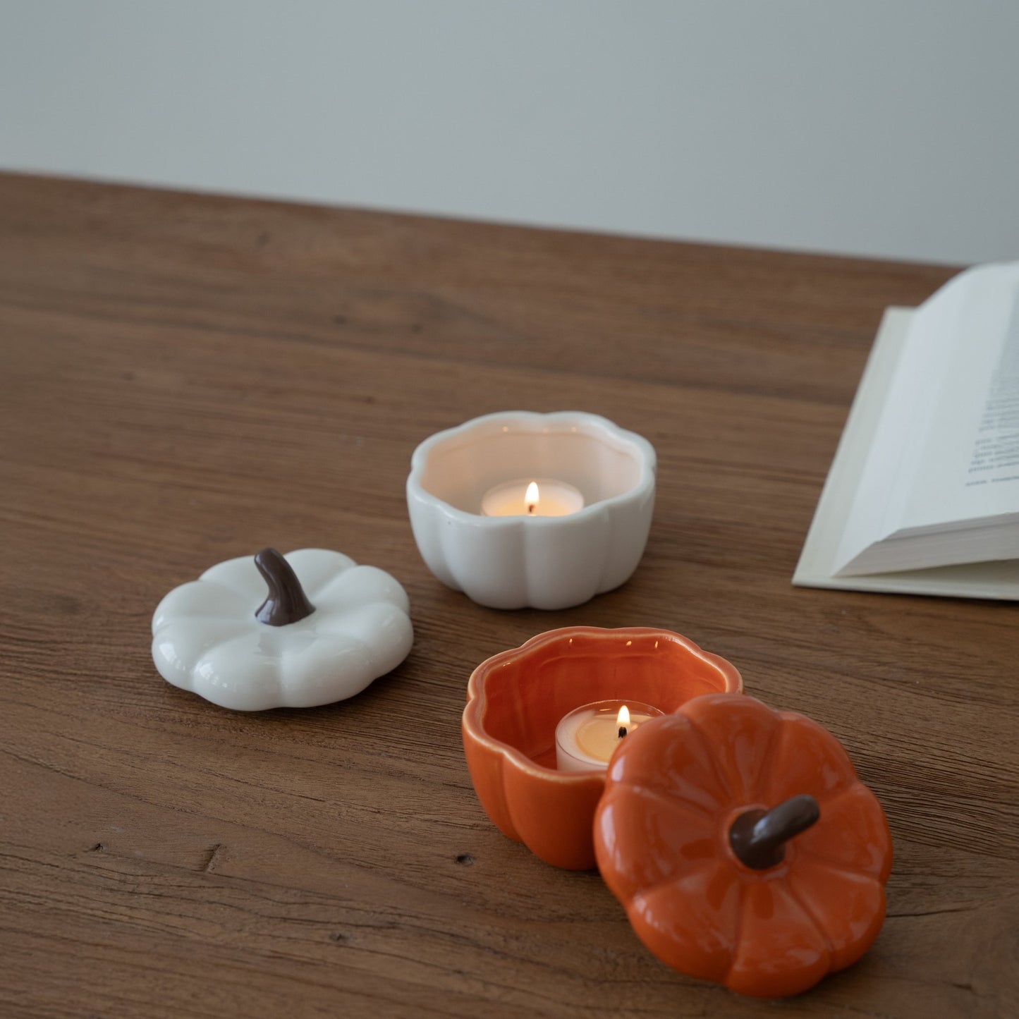 나무 테이블 위에 뚜껑을 열어 티라이트 캔들을 켜둔 주황색 호박 캔들 홀더와 흰색 호박 캔들 홀더, 옆에 펼쳐진 책이 보이는 연출 사진