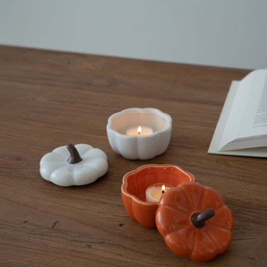 나무 테이블 위에 뚜껑을 열어 티라이트 캔들을 켜둔 주황색 호박 캔들 홀더와 흰색 호박 캔들 홀더, 옆에 펼쳐진 책이 보이는 연출 사진