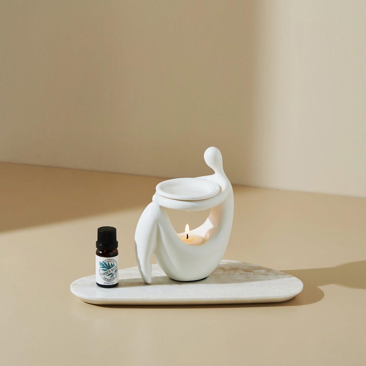 베이지 톤 배경의 테이블 위 타원형 트레이에 올려진 화이트 조형적인 세라믹 오일버너와 불이 켜진 티라이트 캔들, 옆에 세워둔 가든데이즈 에센셜오일 병이 보이는 제품 연출 사진