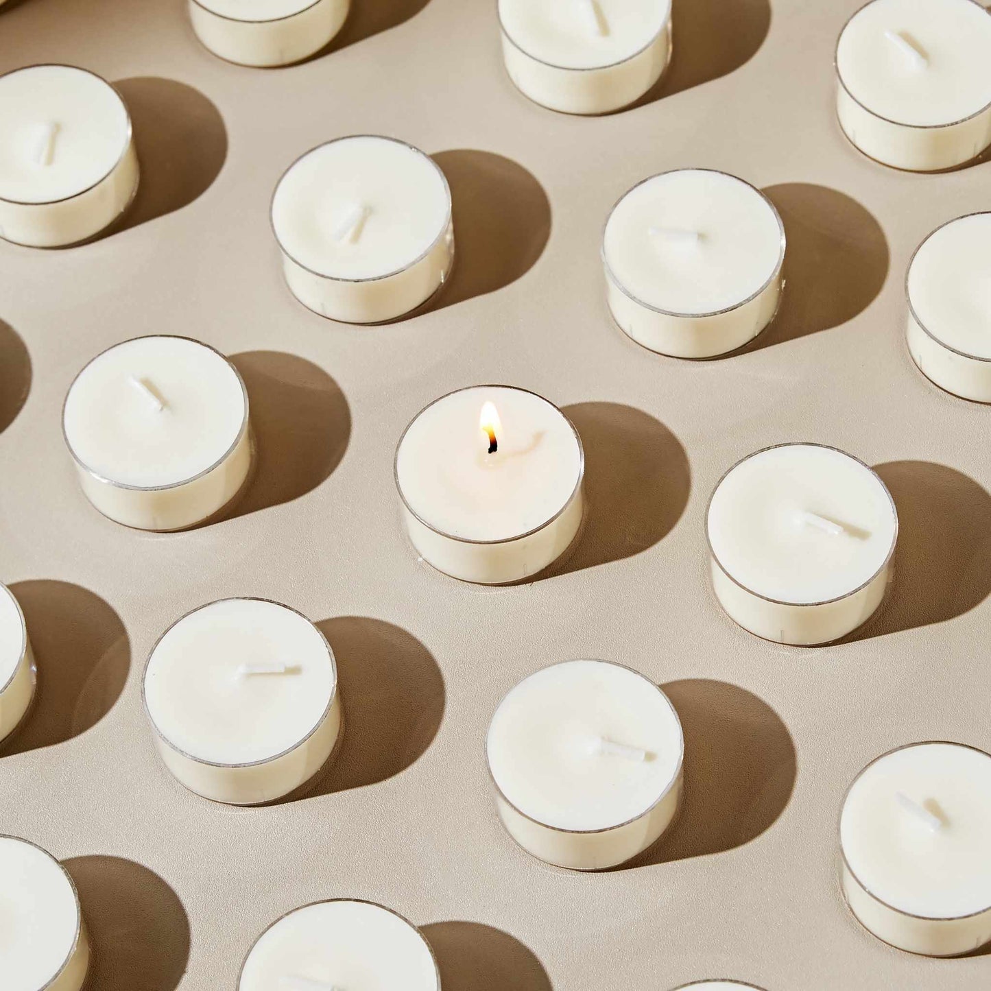 베이지 톤 배경 위에 규칙적으로 배열된 소이 티라이트 캔들들 사이에서 하나만 따로 점등되어 있는 클로즈업 패턴 사진