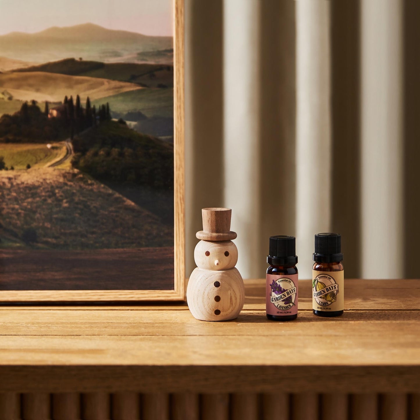 풍경 그림이 있는 나무 선반 위에 우드 스노우맨 디퓨저와 가든데이즈 에센셜오일 병 두 개가 함께 놓인 인테리어 연출 사진