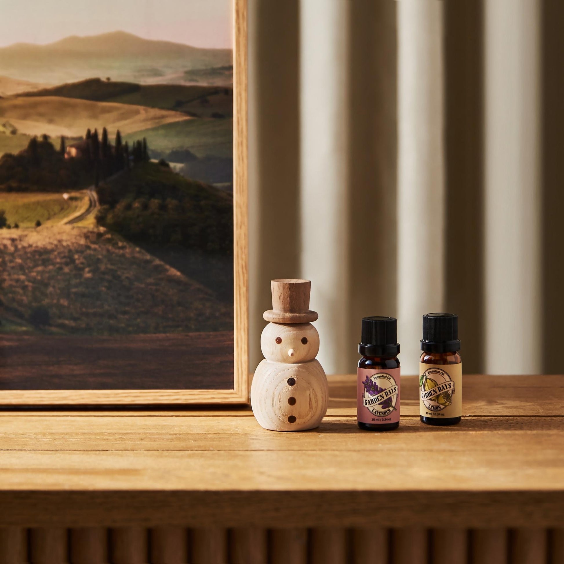 풍경 그림이 있는 나무 선반 위에 우드 스노우맨 디퓨저와 가든데이즈 에센셜오일 병 두 개가 함께 놓인 인테리어 연출 사진