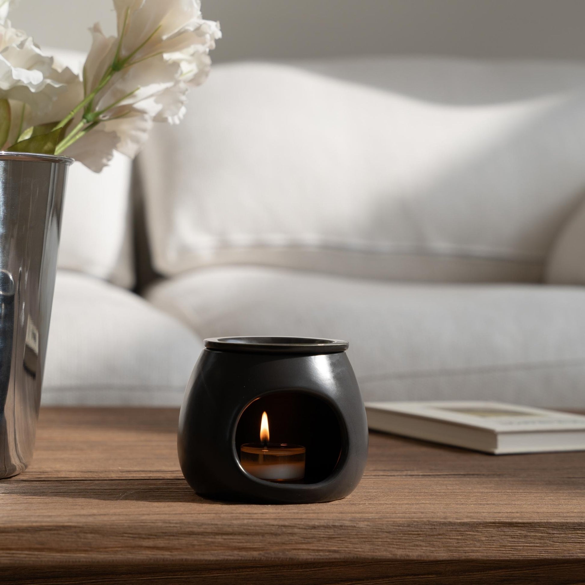 소파 앞 나무 테이블 위 꽃이 담긴 유리병과 책 옆에 놓인 블랙 세라믹 오일버너와 불이 켜진 티라이트 캔들이 보이는 거실 연출 사진