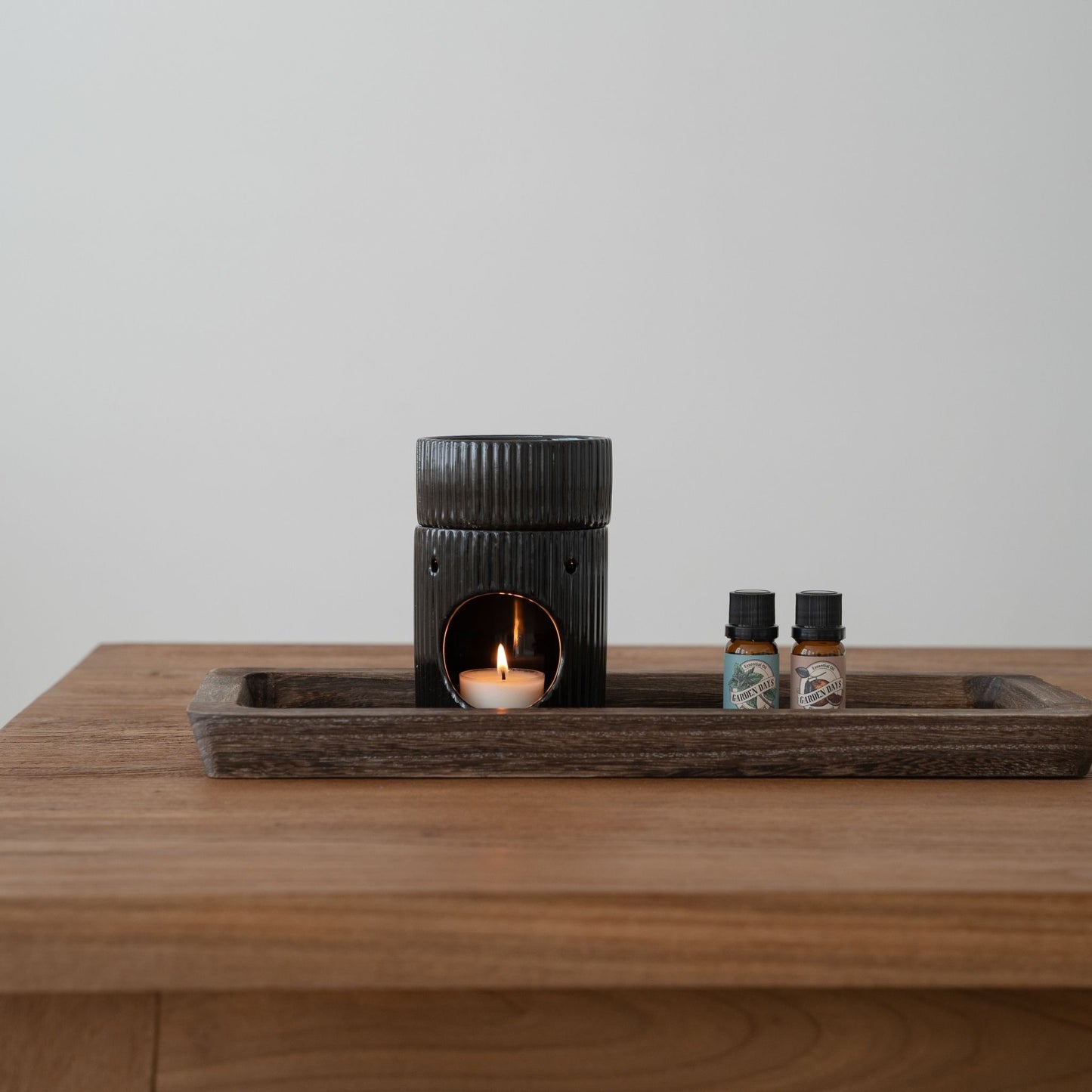 나무 테이블 위 트레이에 올려진 세로 골 패턴 블랙 오일버너와 안에서 은은하게 켜진 티라이트 캔들, 옆에 세워둔 가든데이즈 에센셜오일 병 두 개가 보이는 제품 연출 사진
