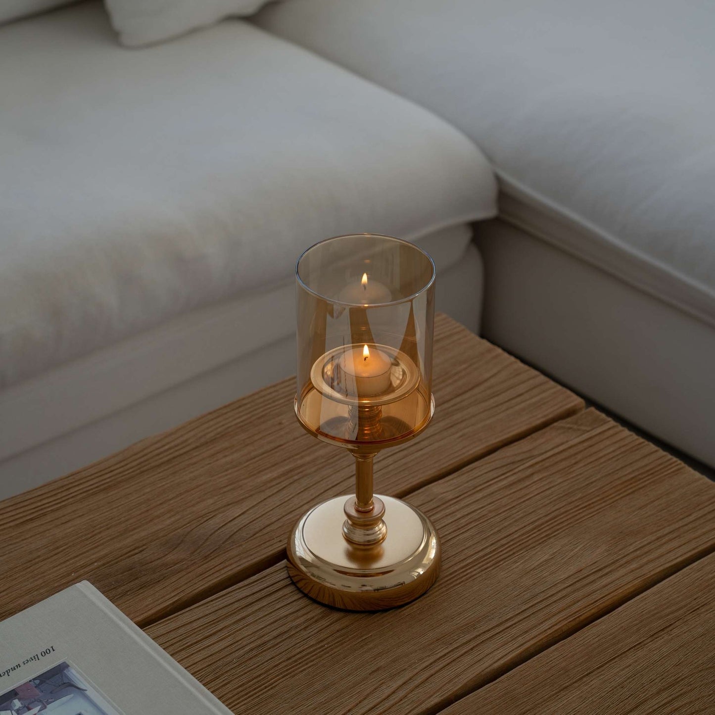 소파 옆 나무 티테이블 위에 올려진 골드 스탠드 유리 캔들 홀더와 따뜻한 불빛의 티라이트 캔들이 보이는 거실 연출 사진