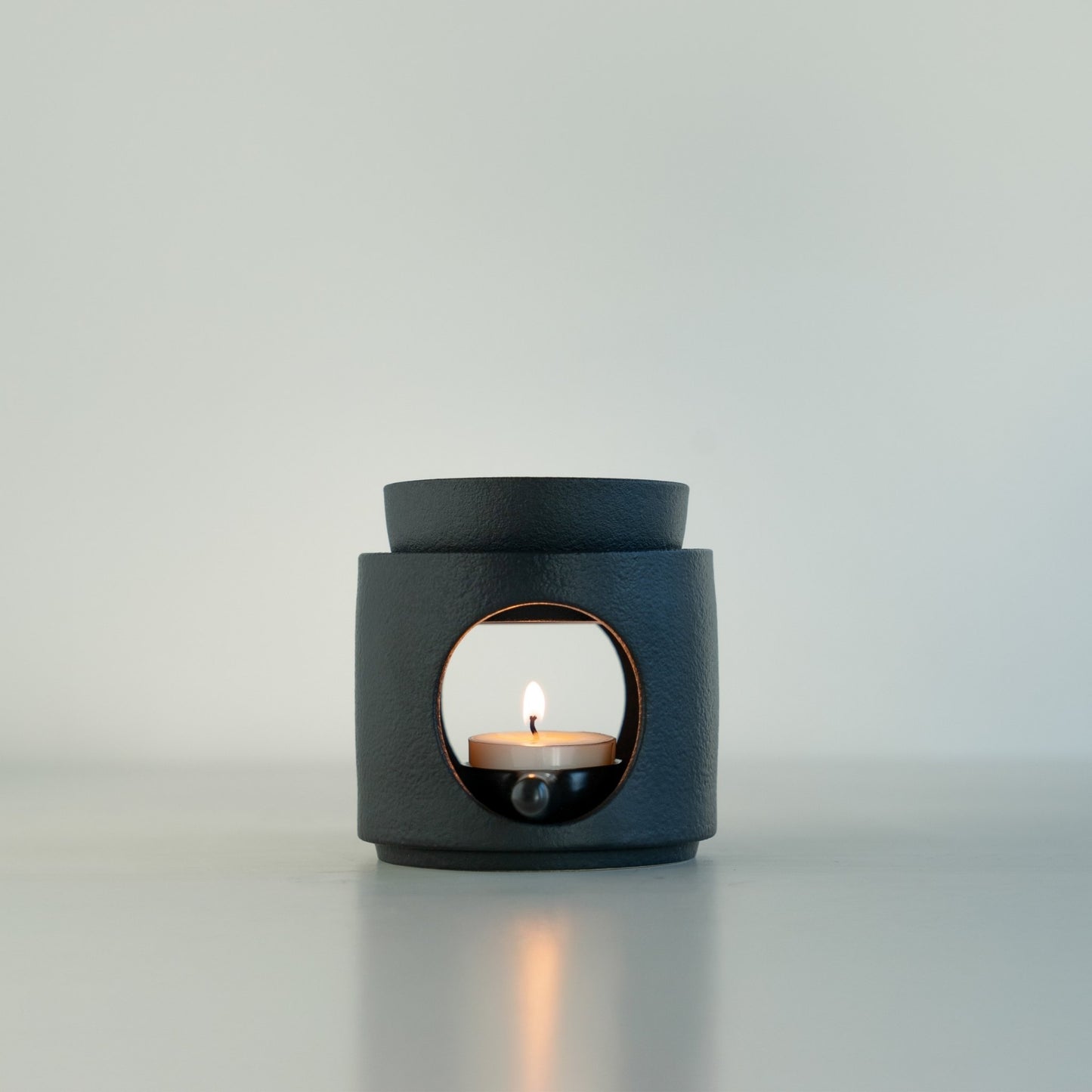 연한 배경 앞 하얀 상판 위에 단독으로 놓인 블랙 세라믹 오일버너와 불이 켜진 티라이트 캔들 정면 사진