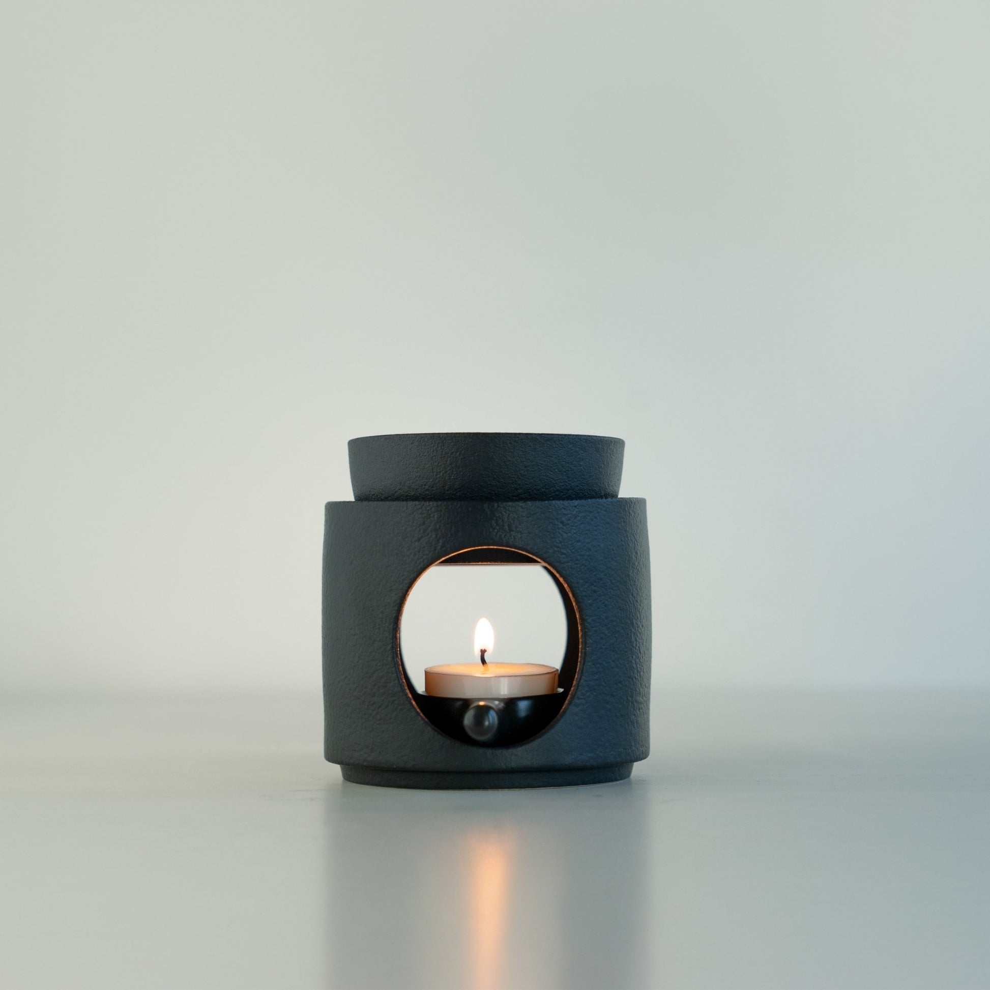 연한 배경 앞 하얀 상판 위에 단독으로 놓인 블랙 세라믹 오일버너와 불이 켜진 티라이트 캔들 정면 사진