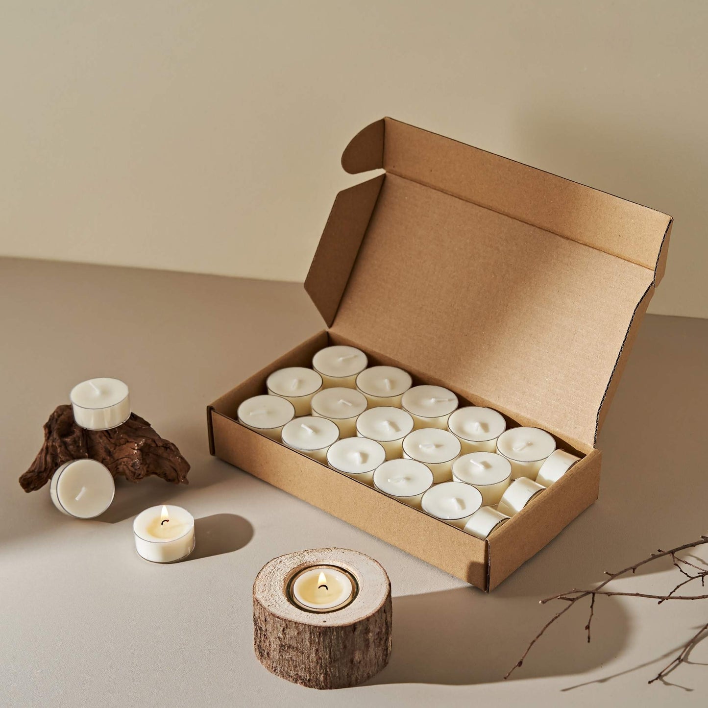 베이지 바닥 위 나무 조각 장식 옆에 놓인 소이 티라이트 캔들 33개입 큰 상자와 상자에서 꺼낸 티라이트 캔들, 앞에 켜져 있는 연출 사진 