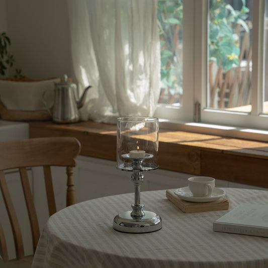 창가가 보이는 다이닝룸에서 줄무늬 테이블보 위에 놓인 실버 스탠드 유리 캔들 홀더와 안에 켜진 티라이트 캔들이 보이는 연출 사진