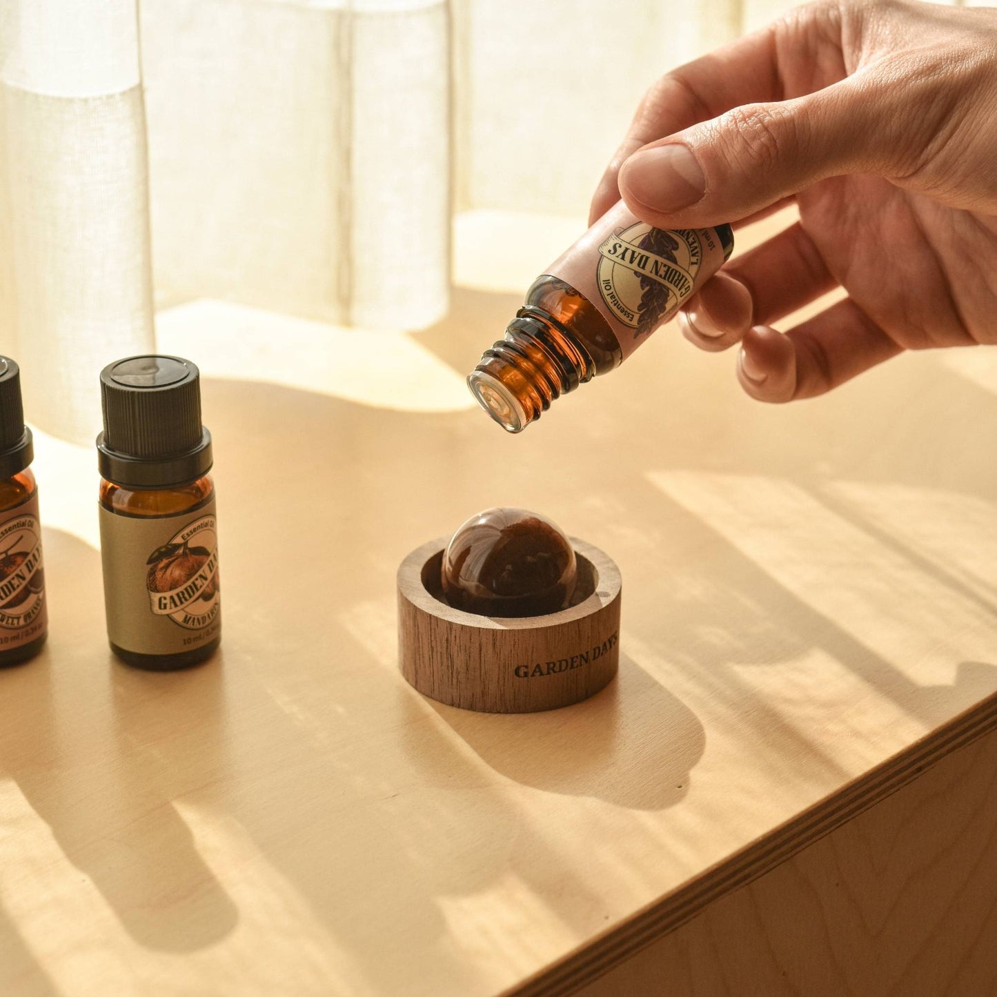 창가 쪽 나무 상판 위에 호두 브리즈볼과 가든데이즈 에센셜오일 병 세 개가 나란히 놓여 있고, 브리즈볼 위로 에센셜오일을 떨어뜨리는 손이 보이는 따뜻한 분위기의 연출 사진