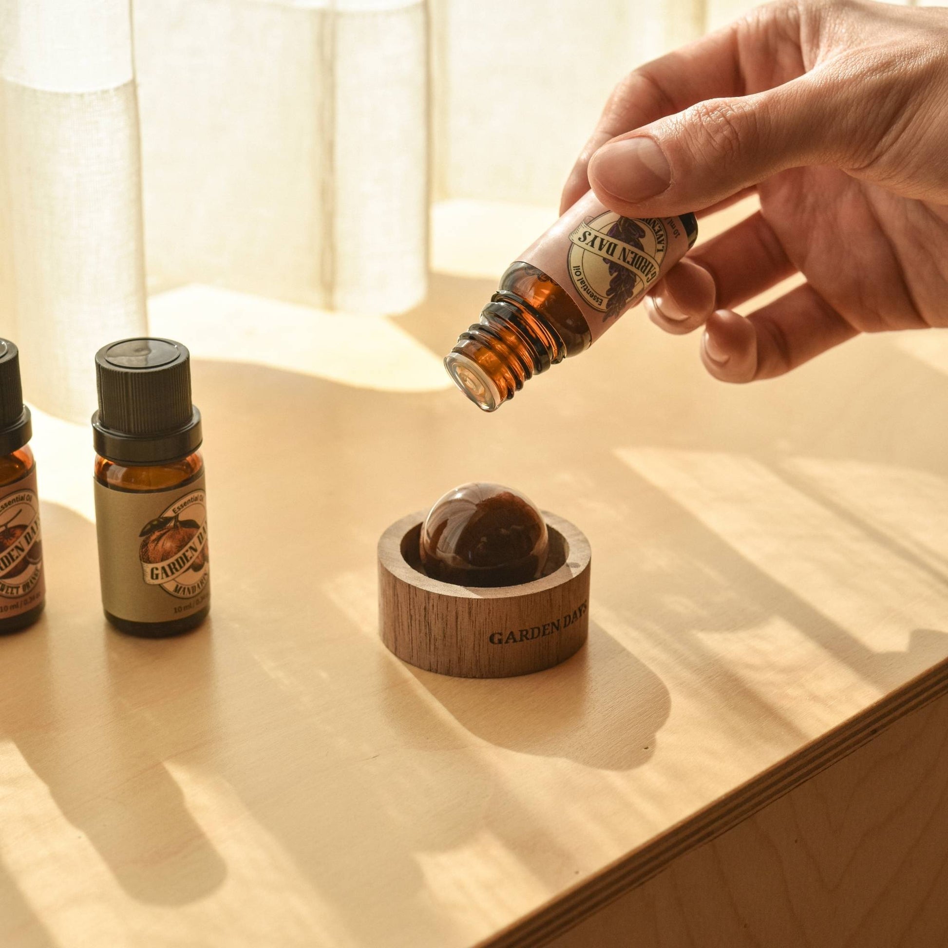창가 쪽 나무 상판 위에 호두 브리즈볼과 가든데이즈 에센셜오일 병 세 개가 나란히 놓여 있고, 브리즈볼 위로 에센셜오일을 떨어뜨리는 손이 보이는 따뜻한 분위기의 연출 사진