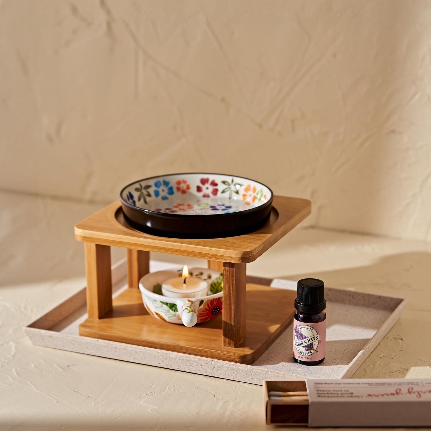 밝은 트레이 위 우드 스탠드 오일버너와 플라워 패턴 세라믹 볼, 아래 불이 켜진 티라이트 캔들, 옆에 세워둔 가든데이즈 에센셜오일 병이 함께 보이는 제품 연출 사진