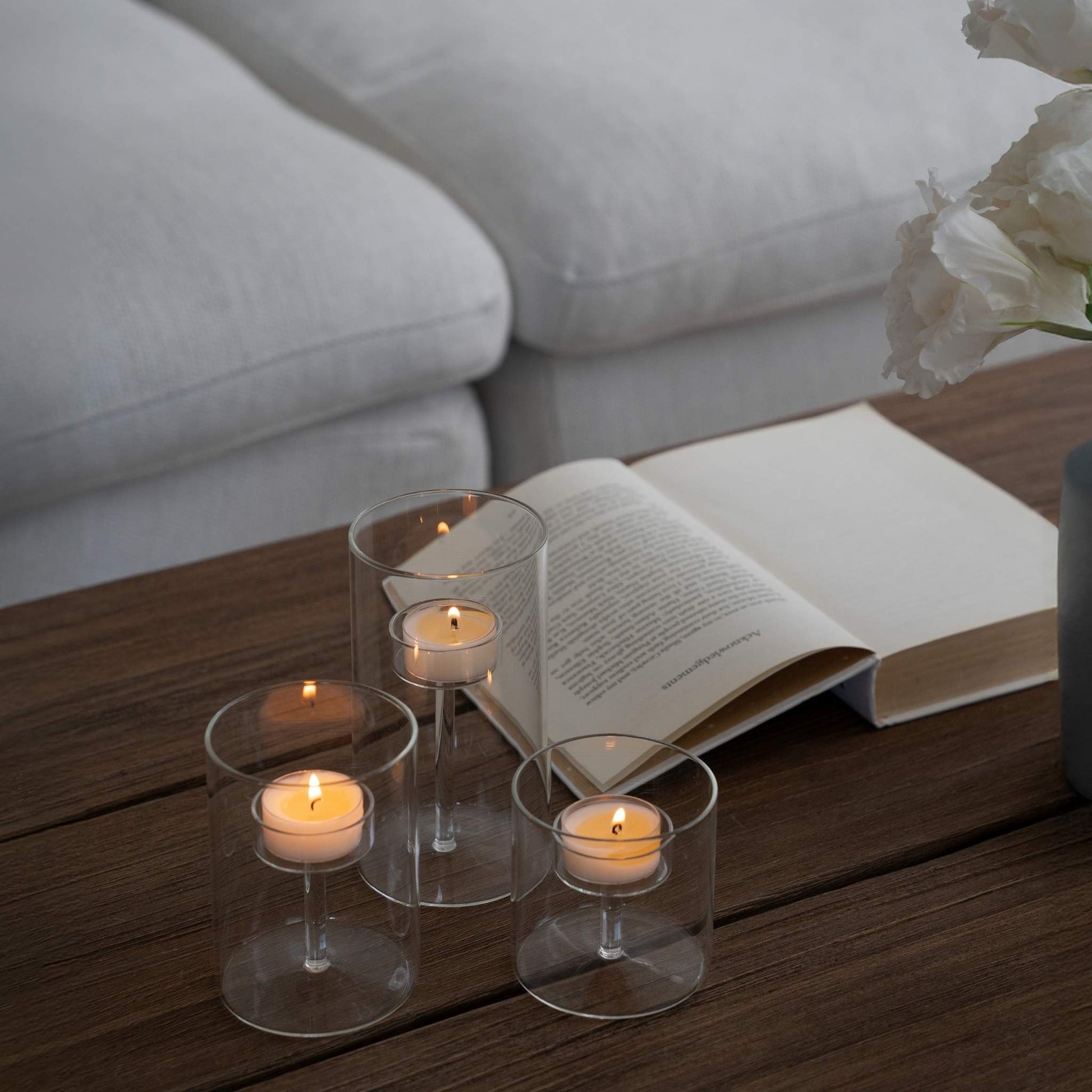 쇼파 앞 테이블에 책이 펼쳐져 있고 투명 유리 티라이트 홀더와 안에서 켜진 티라이트 캔들이 보이는 심플한 연출 사진