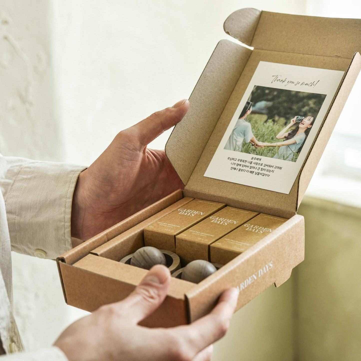 창가 쪽에서 두 손으로 호두 브리즈볼 2개와 에센셜오일 3개가 들어 있는 선물세트 박스를 들고 있는 모습, 뚜껑 안쪽에 맞춤카드가 보이는 사진