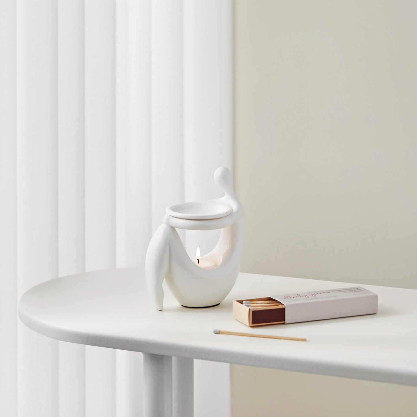 화이트 라운드 테이블 위에 책 더미와 함께 놓인 화이트 조형적인 세라믹 오일버너와 따뜻한 불빛의 티라이트 캔들이 보이는 미니멀 인테리어 연출 사진