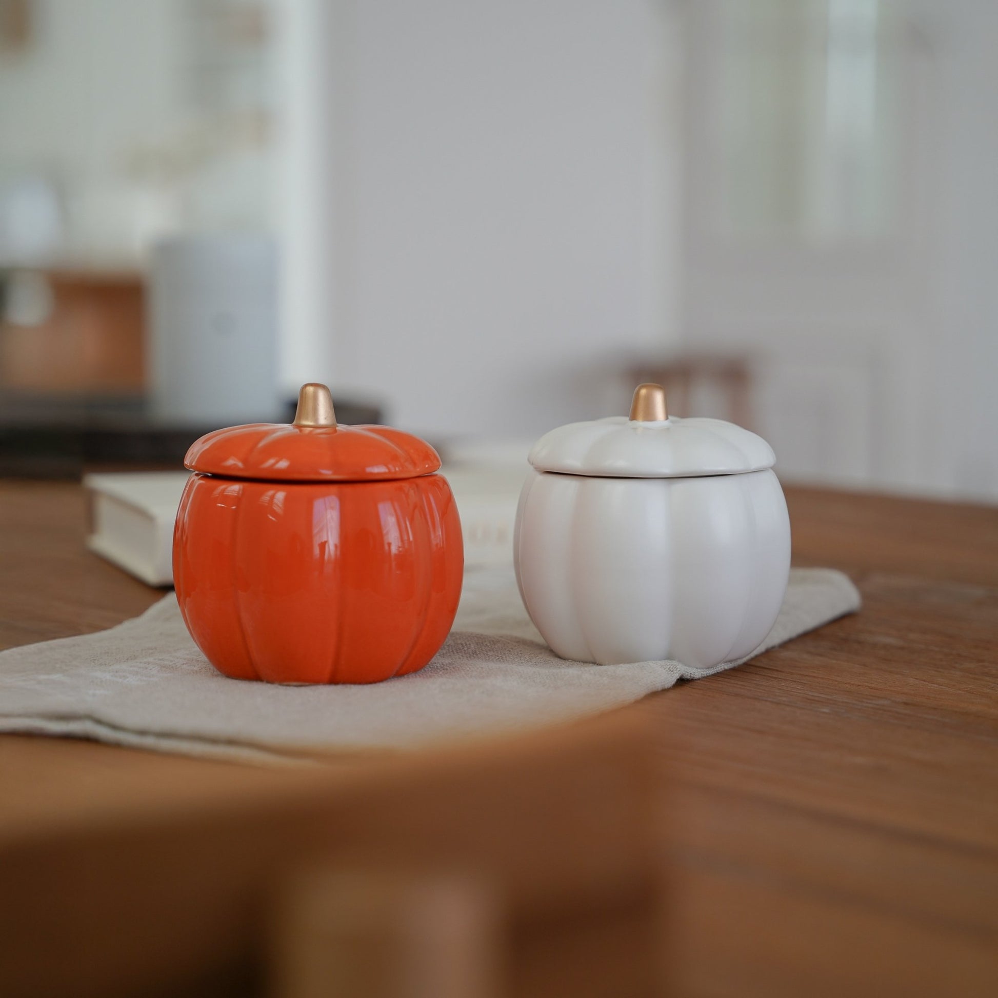 실내가 흐릿하게 보이는 배경 속 식탁 위에 놓인 주황색과 흰색 호박 캔들 홀더 두 개가 있는 가을 인테리어 사진