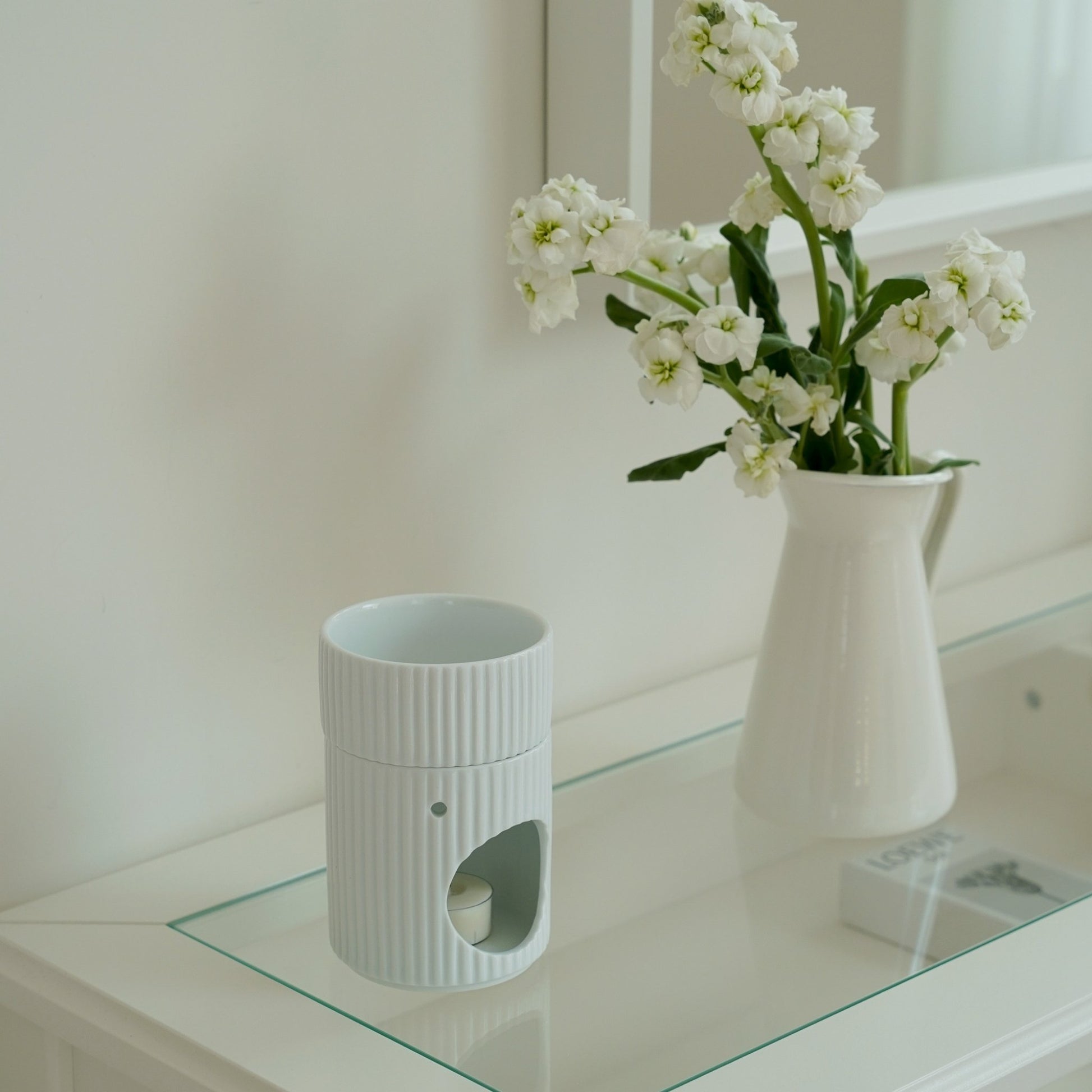 유리 선반 위 꽃이 꽂힌 화이트 화병 옆에 단독으로 놓인 세로 골 패턴 화이트 오일버너와 안쪽이 비어 있는 구조가 보이는 미니멀 인테리어 연출 사진