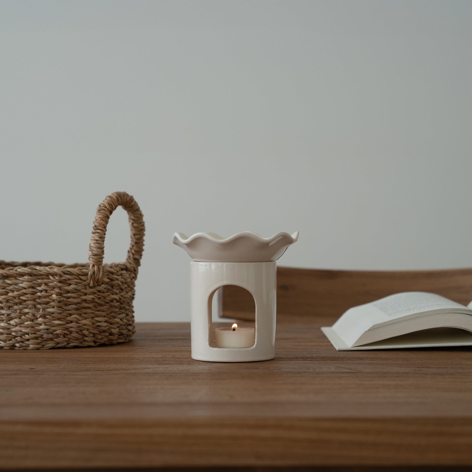 침대 옆 나무 선반 위에 놓인 웨이브  모양 화이트 오일버너와 티라이트 캔들, 옆에 라탄 바스켓과 침구가 보이는 침실 연출 사진