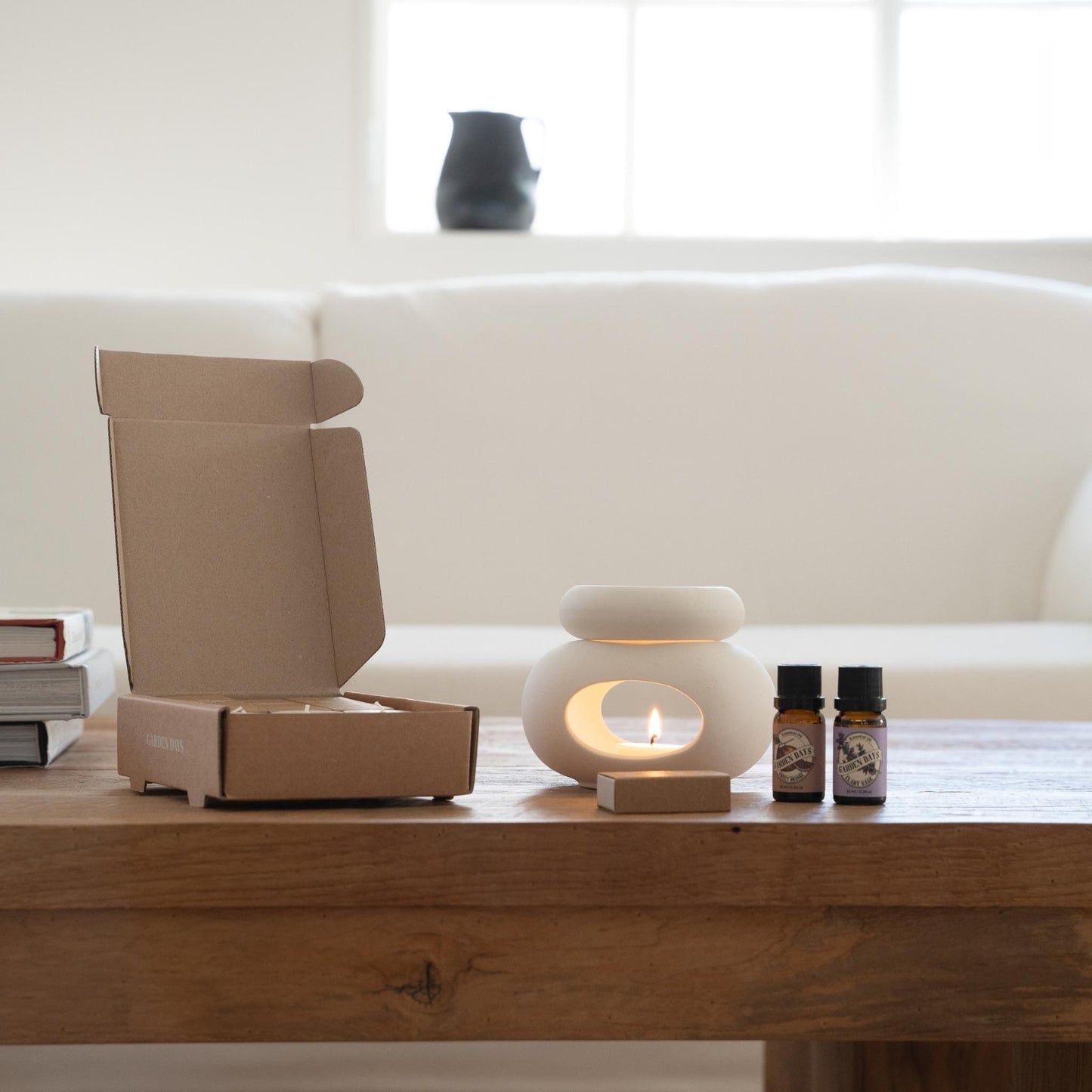 소파 앞 테이블 위에 열린 선물박스 안에 조약돌 오일버너와 구성품이 담겨 있고, 옆에는 닫힌 박스와 에센셜오일 3종이 나란히 놓인 조약돌 오일버너 선물세트 패키지 사진