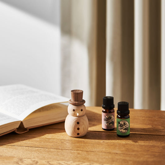 나무 테이블 위 펼쳐진 책 옆에 놓인 우드 스노우맨 디퓨저와 가든데이즈 에센셜오일 병 두 개가 보이는 겨울 무드 연출 사진