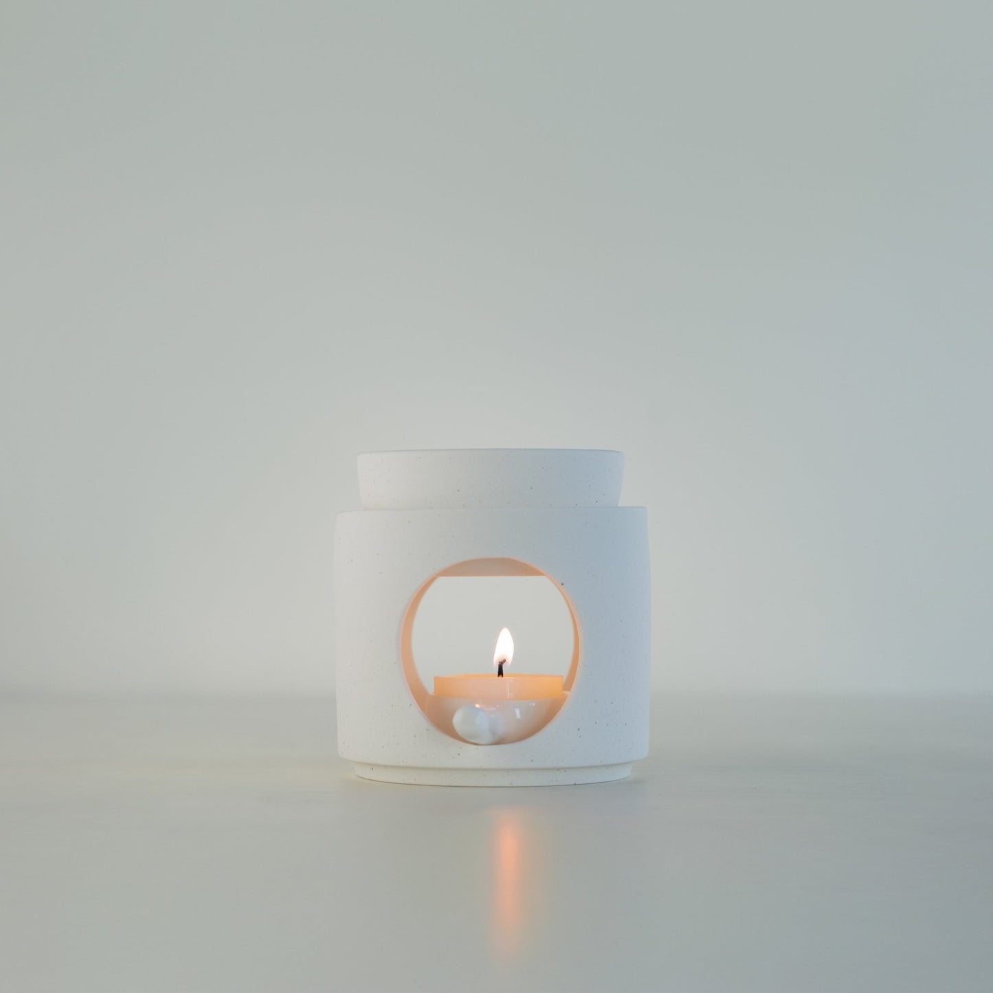 연한 배경 앞 하얀 상판 위에 단독으로 놓인 화이트 세라믹 오일버너와 불이 켜진 티라이트 캔들 정면 사진