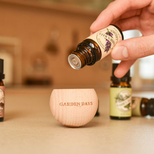 나무 테이블 위 가든데이즈 로고가 새겨진 우드 브리즈볼에 에센셜오일을 떨어뜨리는 손이 보이는 클로즈업 사진