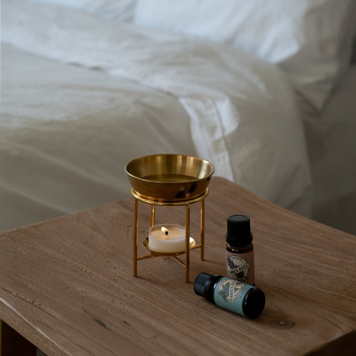 침대 옆 나무 협탁 위에 놓인 골드 스탠드 오일버너와 불이 켜진 티라이트 캔들, 옆에 놓인 가든데이즈 에센셜오일 병 두 개가 보이는 침실 연출 사진
