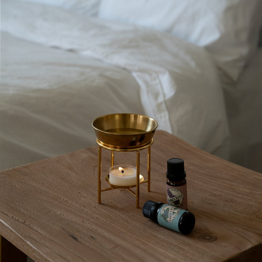 침대 옆 나무 협탁 위에 놓인 골드 스탠드 오일버너와 불이 켜진 티라이트 캔들, 옆에 놓인 가든데이즈 에센셜오일 병 두 개가 보이는 침실 연출 사진