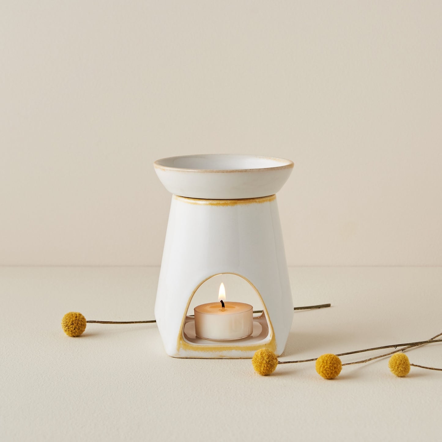 크림색 배경 위 노란 가장자리가 포인트인 화이트 세라믹 옐로우 오일버너와 불이 켜진 티라이트 캔들, 옆에 놓인 노란 꽃 장식이 보이는 제품 연출 사진