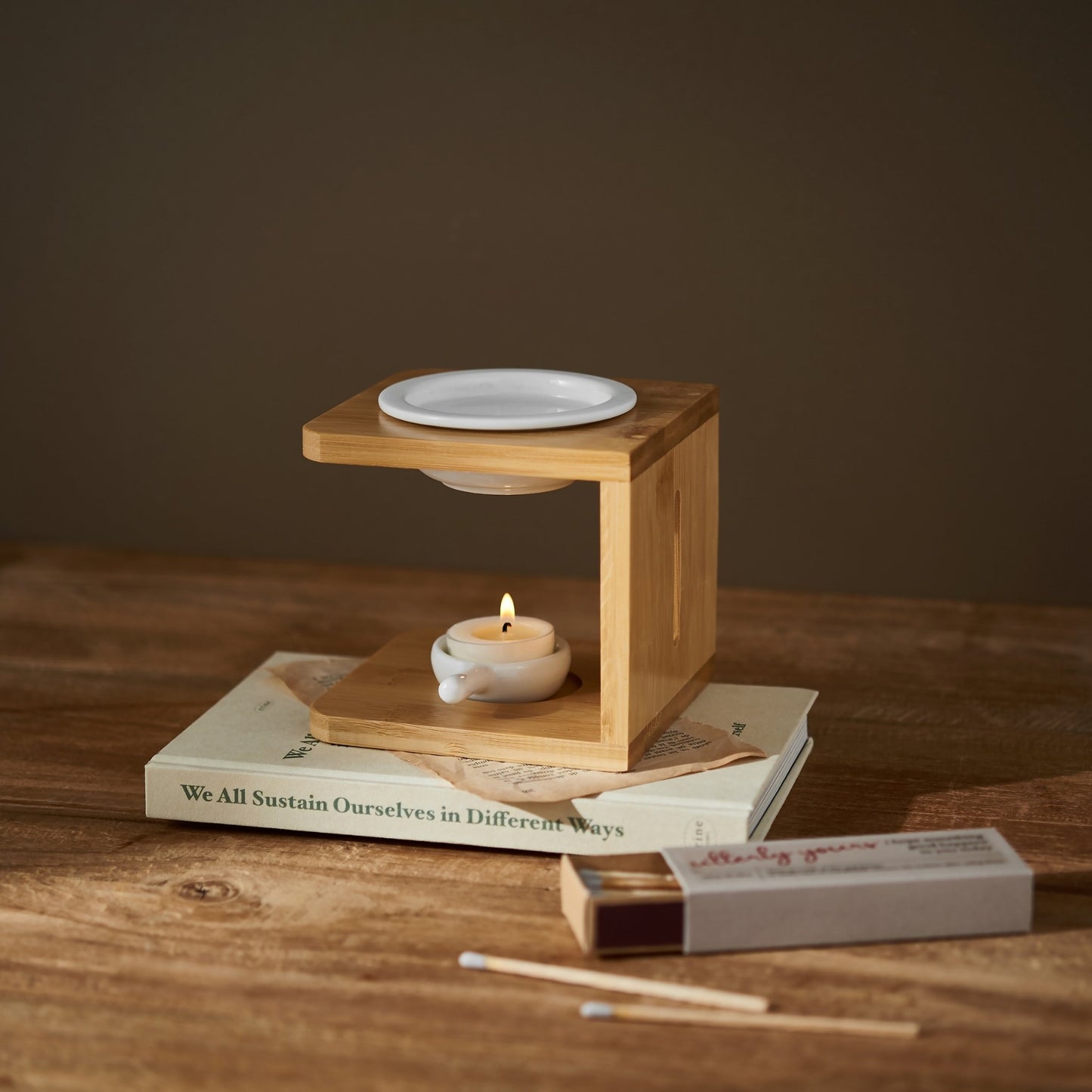 책 위에 올려진 우드 오일버너와 아래쪽 불이 켜진 티라이트 캔들, 옆에 놓인 성냥갑이 보이는 따뜻한 분위기의 연출 사진