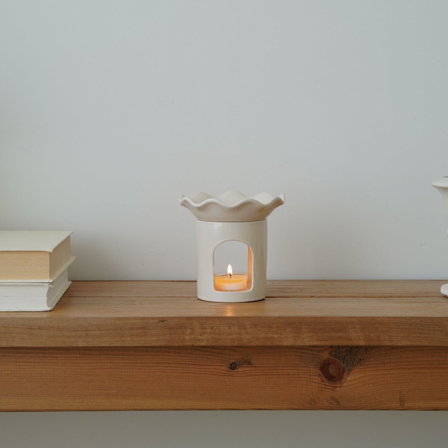 나무 선반 위 책 더미 옆에 놓인 웨이브  모양 화이트 세라믹 오일버너와 안에서 켜진 티라이트 캔들이 보이는 메인 연출 사진