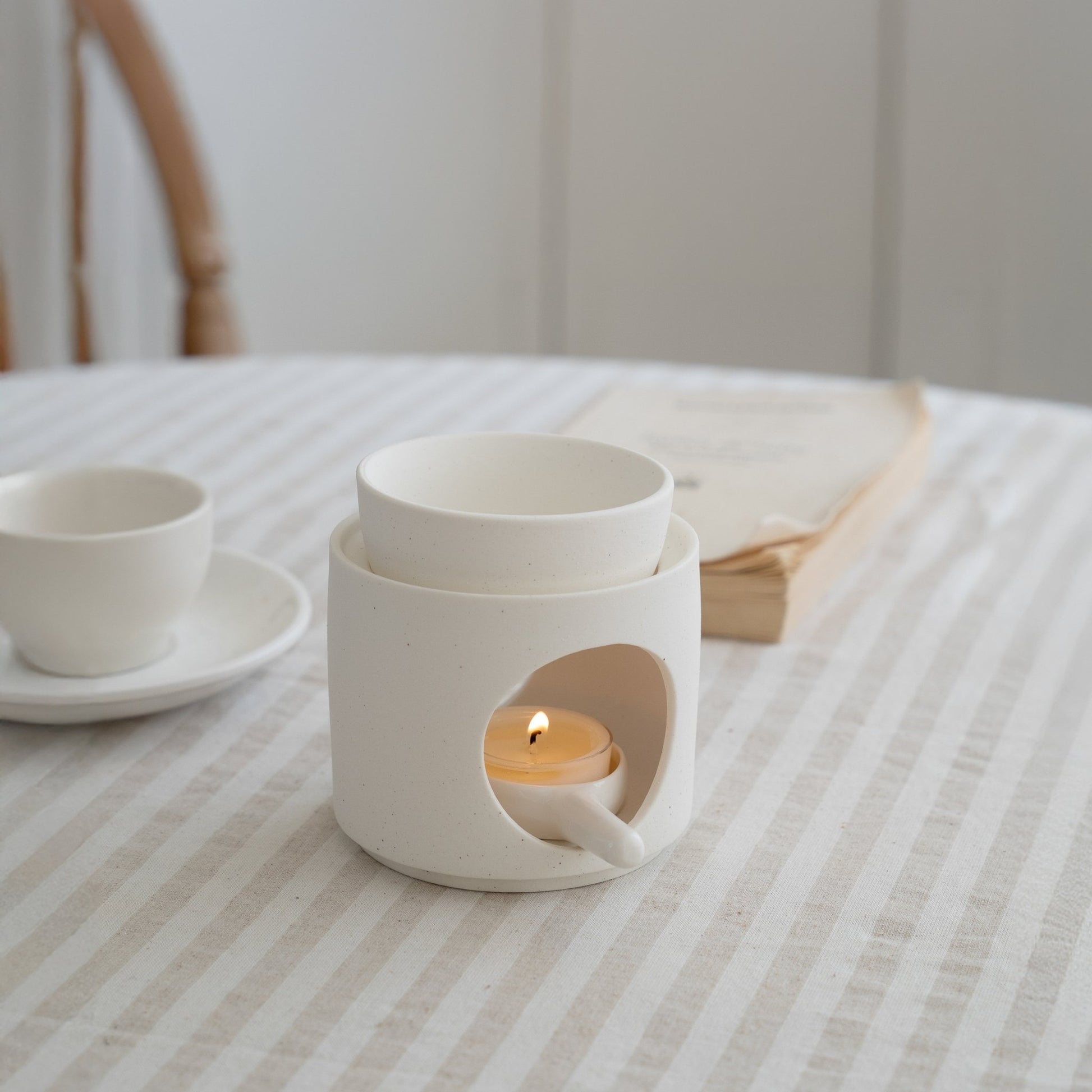 줄무늬 테이블보 위에 책과 찻잔 옆에 놓인 화이트 세라믹 오일버너와 안에서 켜진 티라이트 캔들이 보이는 연출 사진