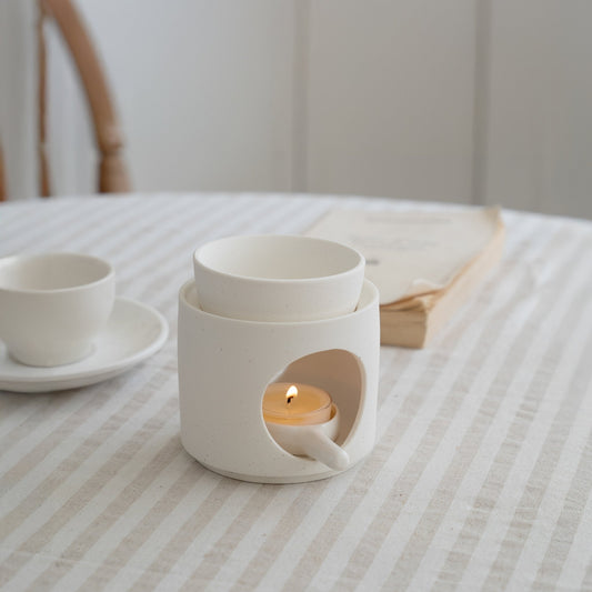 줄무늬 테이블보 위에 책과 찻잔 옆에 놓인 화이트 세라믹 오일버너와 안에서 켜진 티라이트 캔들이 보이는 연출 사진