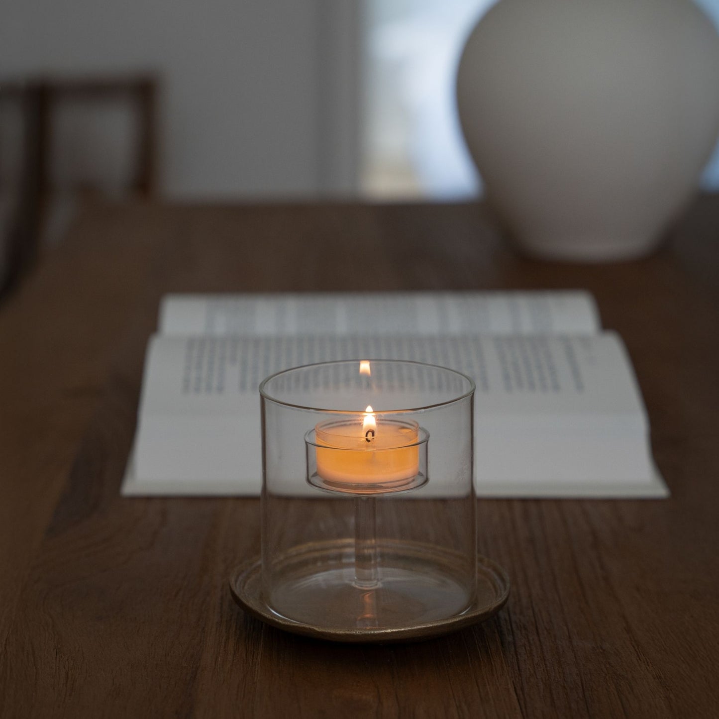 부드러운 조명 아래 접시 위에 단독으로 놓인 투명 유리 티라이트 홀더와 안에서 켜진 티라이트 캔들이 보이는 심플한 연출 사진