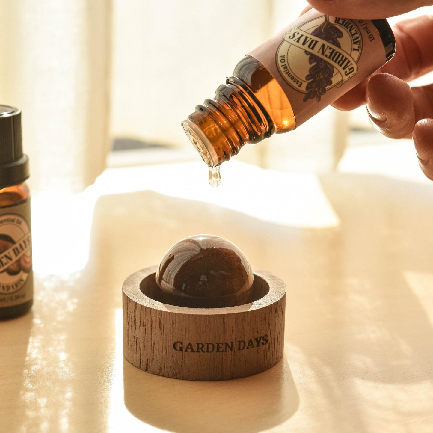 밝은 배경에서 호두 브리즈볼 위로 에센셜오일을 떨어뜨리는 손과 옆에 놓인 가든데이즈 에센셜오일 병들이 보이는 클로즈업 사용 연출 사진