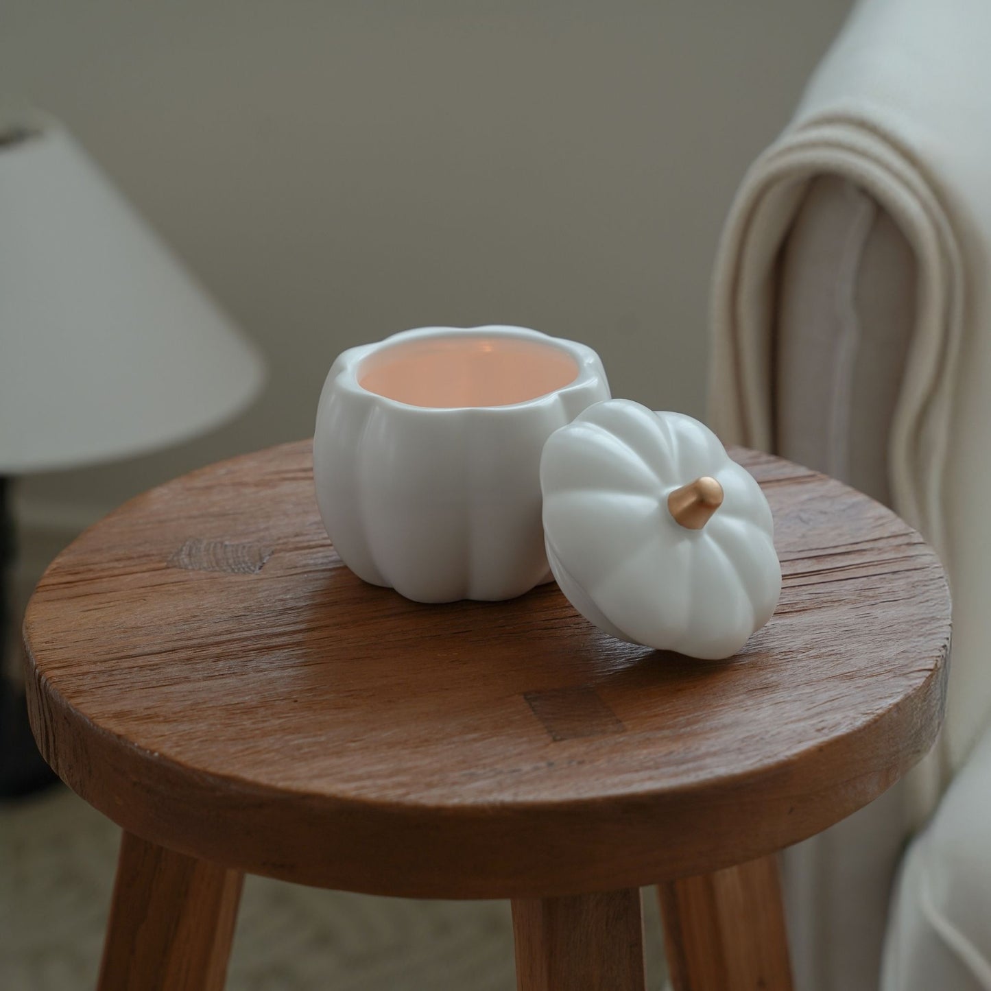 라운드 우드 스툴 위에 놓인 흰색 호박 캔들 홀더와 뚜껑, 뒤로 의자와 쿠션이 보이는 아늑한 연출 사진