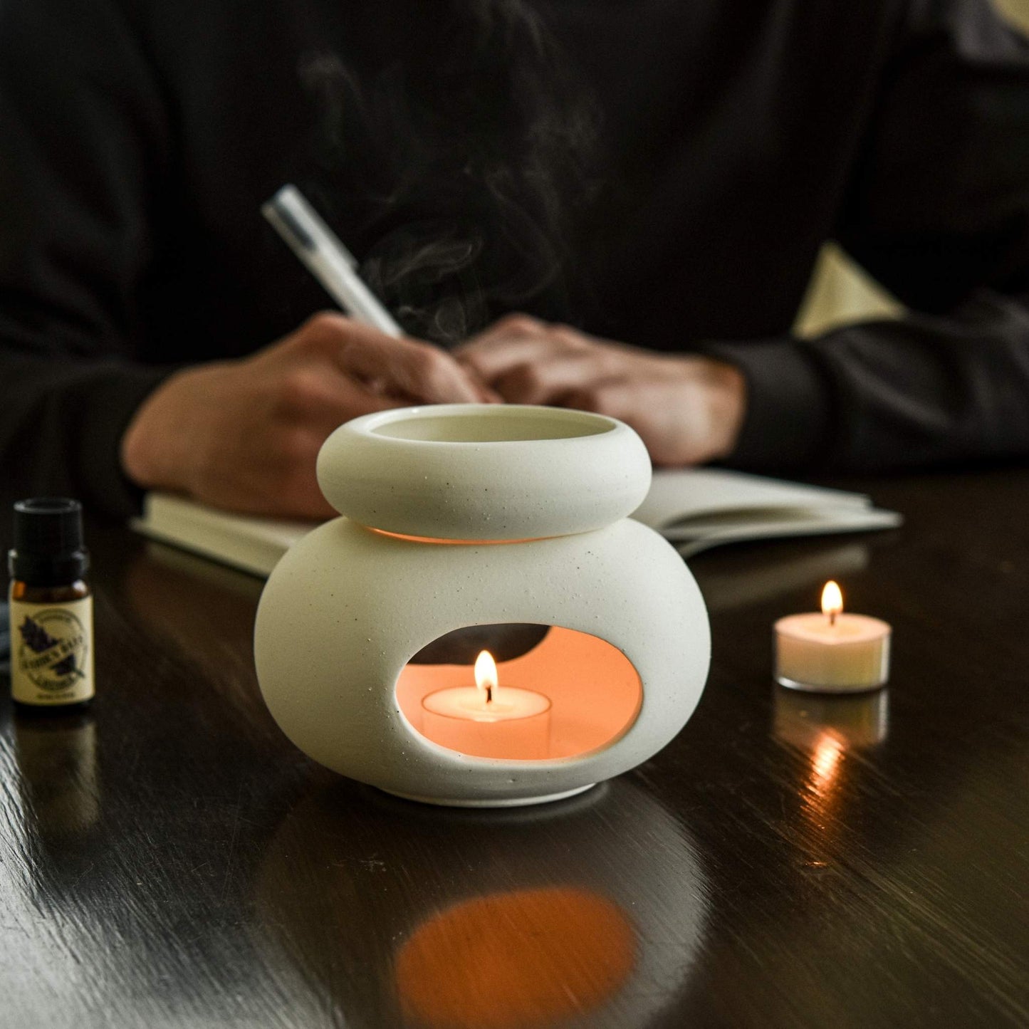 어두운 톤 책상 위에서 노트를 쓰고 있는 사람 옆으로, 세라믹 오일버너 안에서 타는 소이 티라이트 캔들과 옆에 놓인 캔들·에센셜오일 병이 보이는 사용 연출 사진