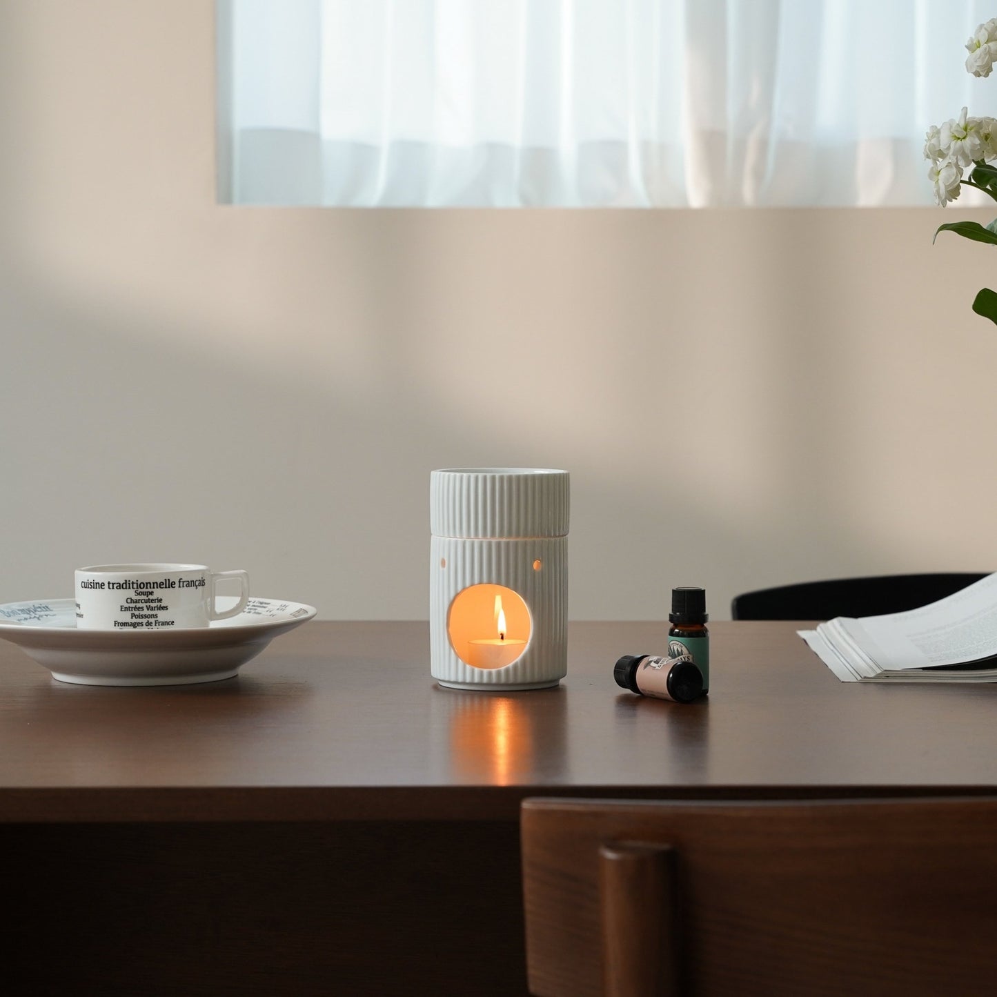 창가가 보이는 책상 위 컵과 소서, 노트 옆에 세워진 세로 골 패턴 화이트 오일버너와 따뜻한 불빛의 티라이트 캔들이 보이는 작업 공간 연출 사진